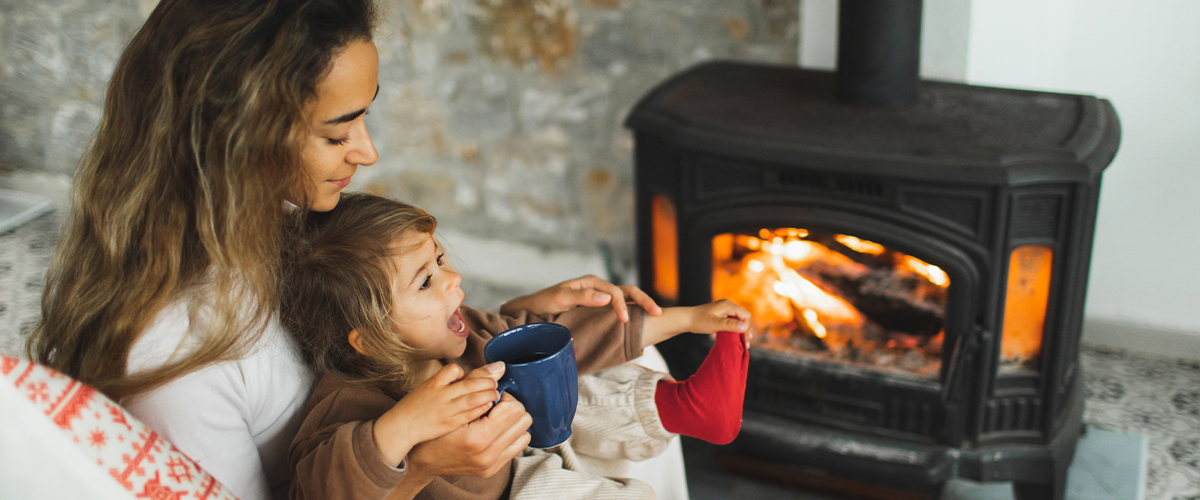 Woman holds a small child while sipping from a mug in front of a fire.