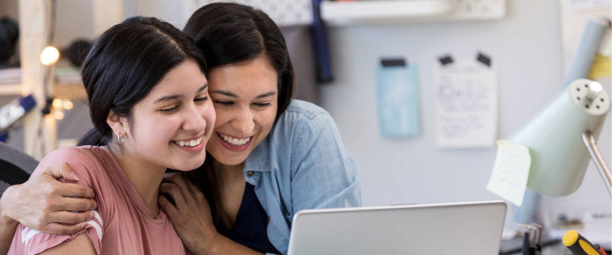 A parent and teenage child hugging while looking at a computer screen.