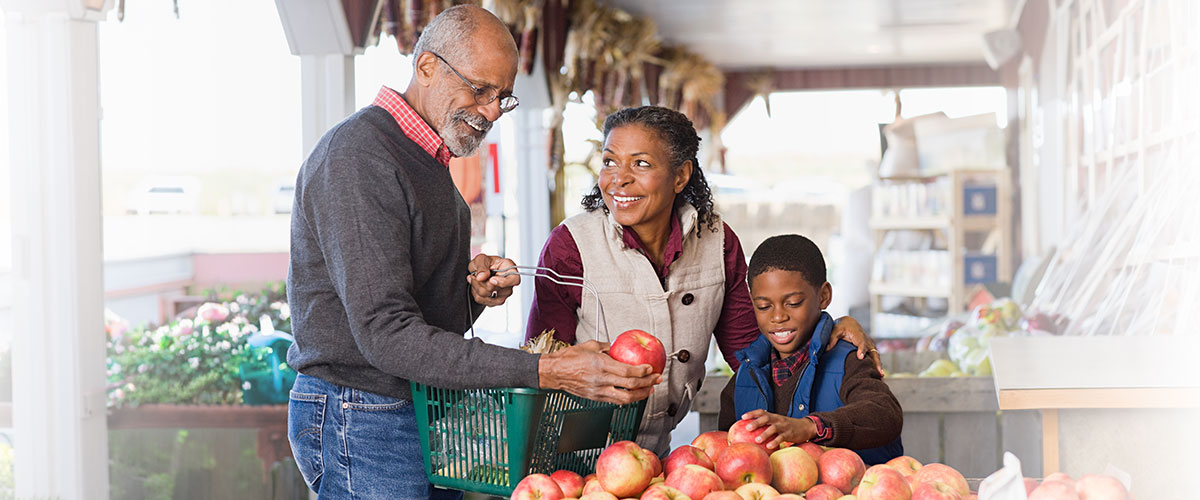 Three people shopping together at a market, selecting fresh produce and placing items in a basket.