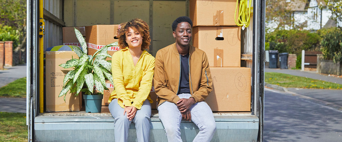 A young couple seated on the back of a moving truck, surrounded by moving boxes.