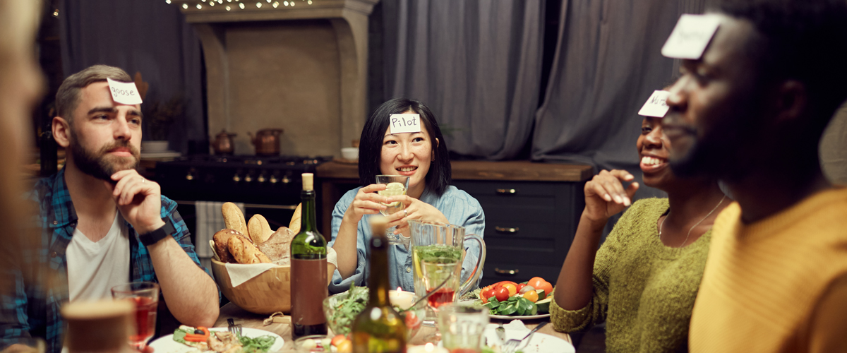 Friends sitting around a table playing a game with playing cards on their head.