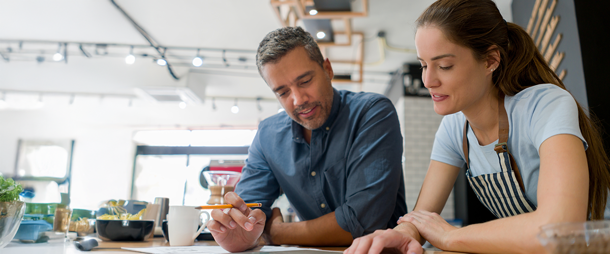 Two coworkers collaborate at a café-style workspace, reviewing notes on a tablet and notebook while planning together in a bright, creative environment.