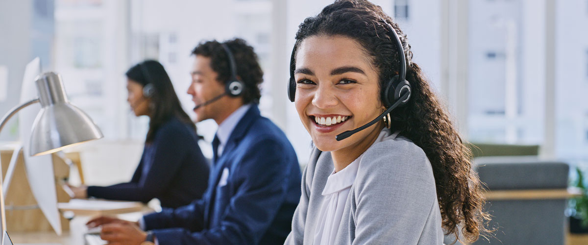 Woman in a customer service setting, smiling while sitting at a desk and wearing a headset.