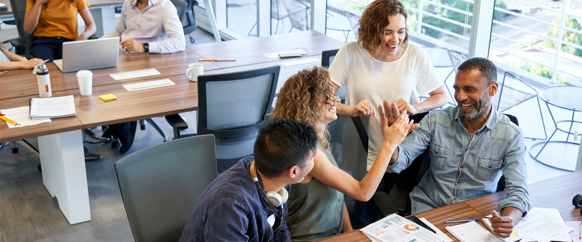 A diverse group of coworkers smile and high-five around a tablet at a shared table, celebrating a collaborative success in a bright, modern office.
