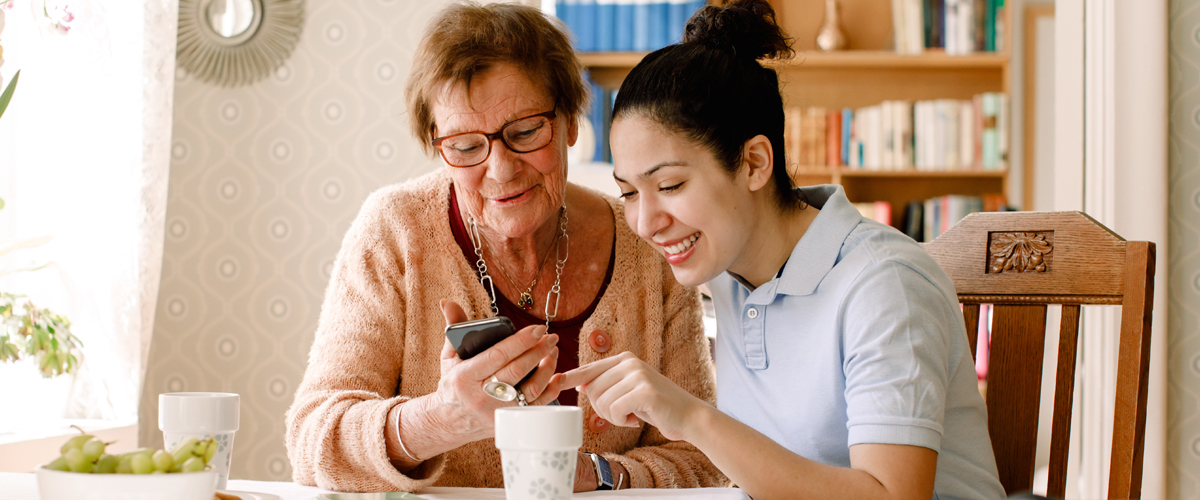 Woman helping a senior use her phone while sitting at a kitchen table.