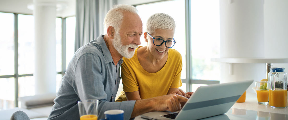 An older couple sits together at a table, smiling and looking at a tablet while enjoying breakfast, sharing a relaxed moment of connection and everyday independence at home.