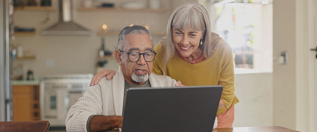 Senior couple sitting at a kitchen table looking at a laptop