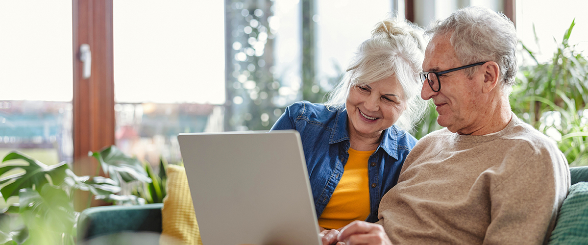 Two adults sitting together on a couch at home, looking at a laptop computer in a bright living room with large windows and indoor plants. The image represents online health resources, retirement planning, digital healthcare access, insurance information, telehealth services, and managing personal finances or benefits from home.