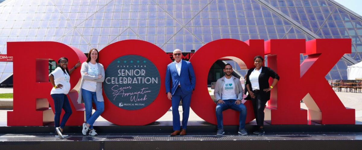 Group of older adults pose smiling in front of the large red ROCK sign outside the Rock and Roll Hall of Fame, celebrating a Senior Celebration event together.
