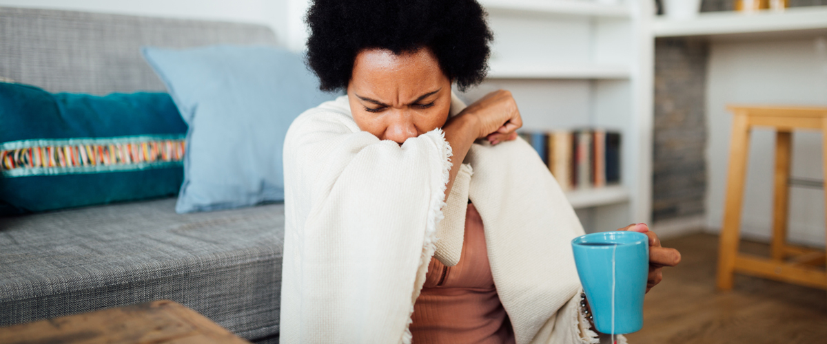 Woman sitting on the floor, holding a mug filled with tea and coughing into her elbow.