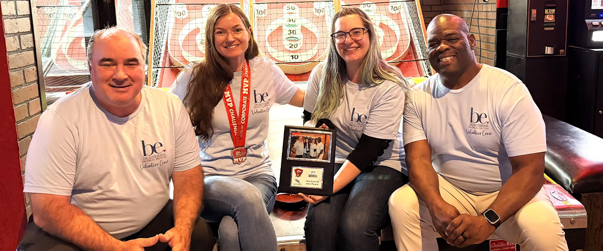 Four employees wearing matching Medical Mutual volunteer shirts sit together in front of arcade skee-ball lanes, holding a Cleveland Corporate Challenge plaque and medal while posing for a group photo.