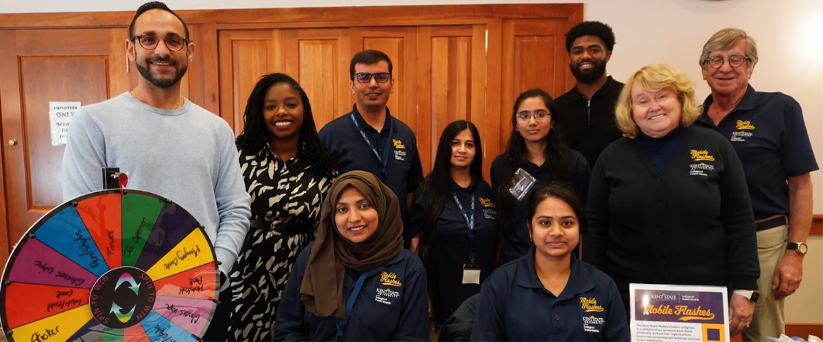 Group photo of eight team members standing and seated indoors in front of wooden doors, wearing casual and branded attire.