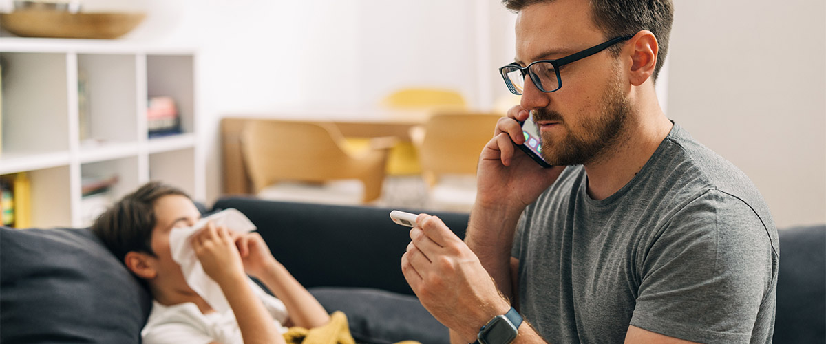 Person speaking on a phone while holding a thermometer on a couch, with another person resting in the background of a living room.