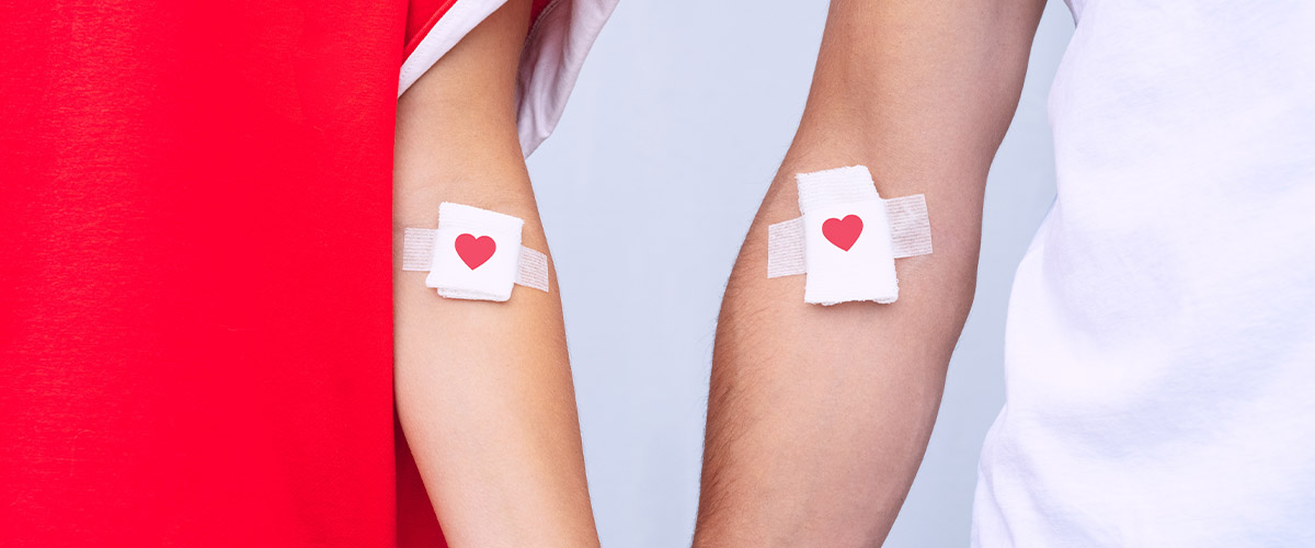 Person’s forearms shown side by side with small bandages placed at the inner elbows, each featuring a red heart icon, suggesting blood donation; set against a split red and light gray background.