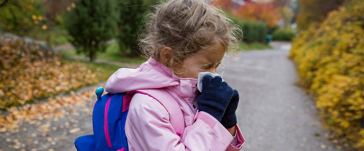 Child in a pink jacket and blue gloves holding a tissue to nose while walking on a tree-lined path covered with autumn leaves.