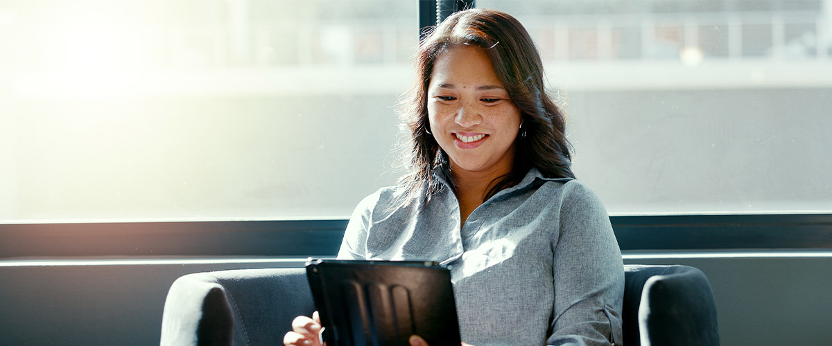 Smiling woman sitting near a large window with bright sunlight, using a tablet device in a modern office setting. The image conveys productivity, digital engagement, and professional confidence in a bright workspace.