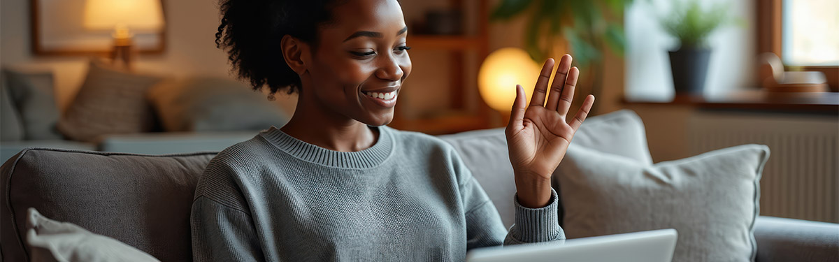 Person sitting on a couch using a laptop, raising one hand as if in a video call. The setting appears to be a softly lit living room with a lamp, plants, and a window in the background.