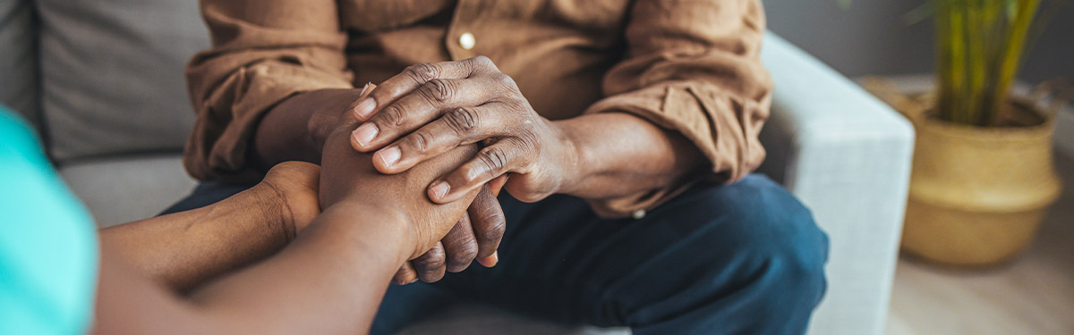 Two people sitting closely as one person gently holds both hands of the other, shown from the forearms down. They are seated on a couch in an indoor setting with a plant in the background.