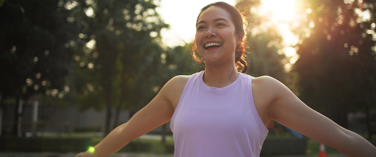 Person in a light athletic top outdoors, illuminated by warm sunlight with trees in the background.