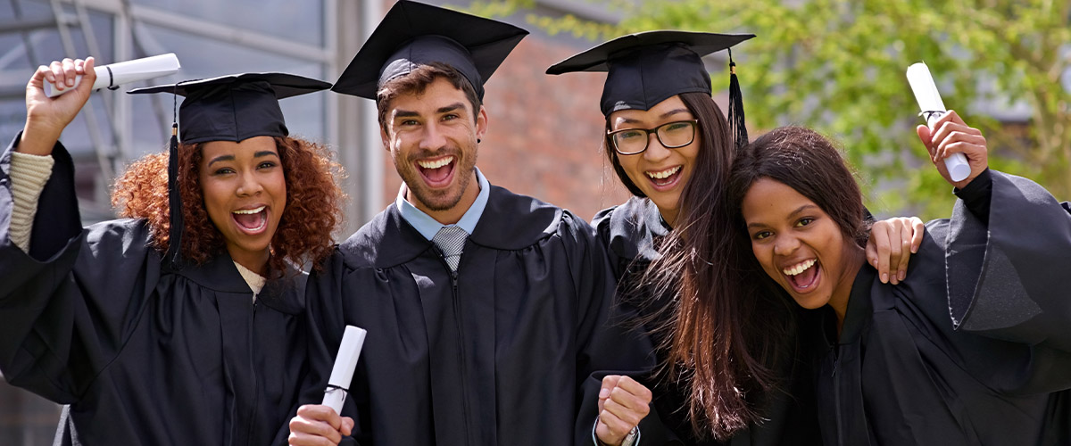 Four graduates in caps and gowns stand together outdoors, holding diplomas and posing for a graduation celebration photo.