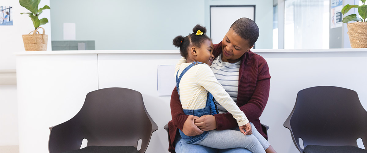 Person holding a child while seated in a healthcare waiting room with empty chairs and a reception desk in the background.