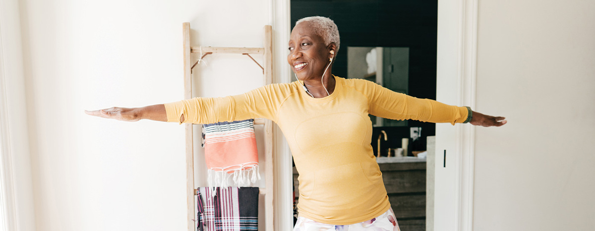 A person stands in a bright room with arms extended outward, practicing a balance or stretching pose near a shelf holding neatly folded towels.