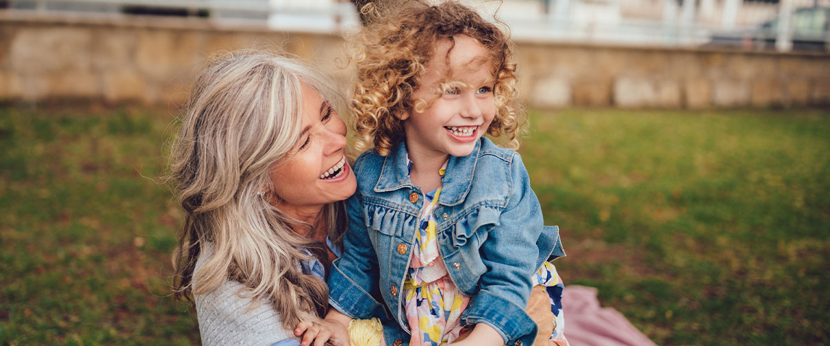 Woman and a child smiling, sitting in the grass outside.